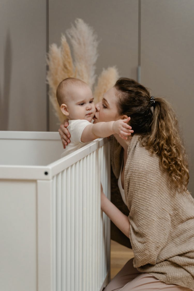 A Woman In Knitted Sweater Kissing Her Baby On The Crib