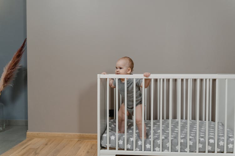 A Baby In Gray Baby Onesie Standing In A Crib