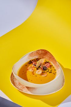 Stylish presentation of pumpkin soup with shrimp and herbs in a ceramic bowl on a vibrant yellow backdrop.