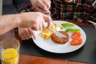 Person Holding Fork and Knife Slicing Meat on White Ceramic Plate