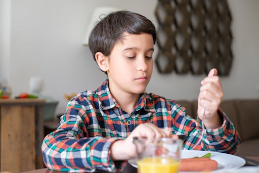 A young boy in a plaid shirt enjoys breakfast with juice and utensils at a home table.