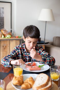A child in a plaid shirt enjoys breakfast with orange juice, croissants, and bread at home.