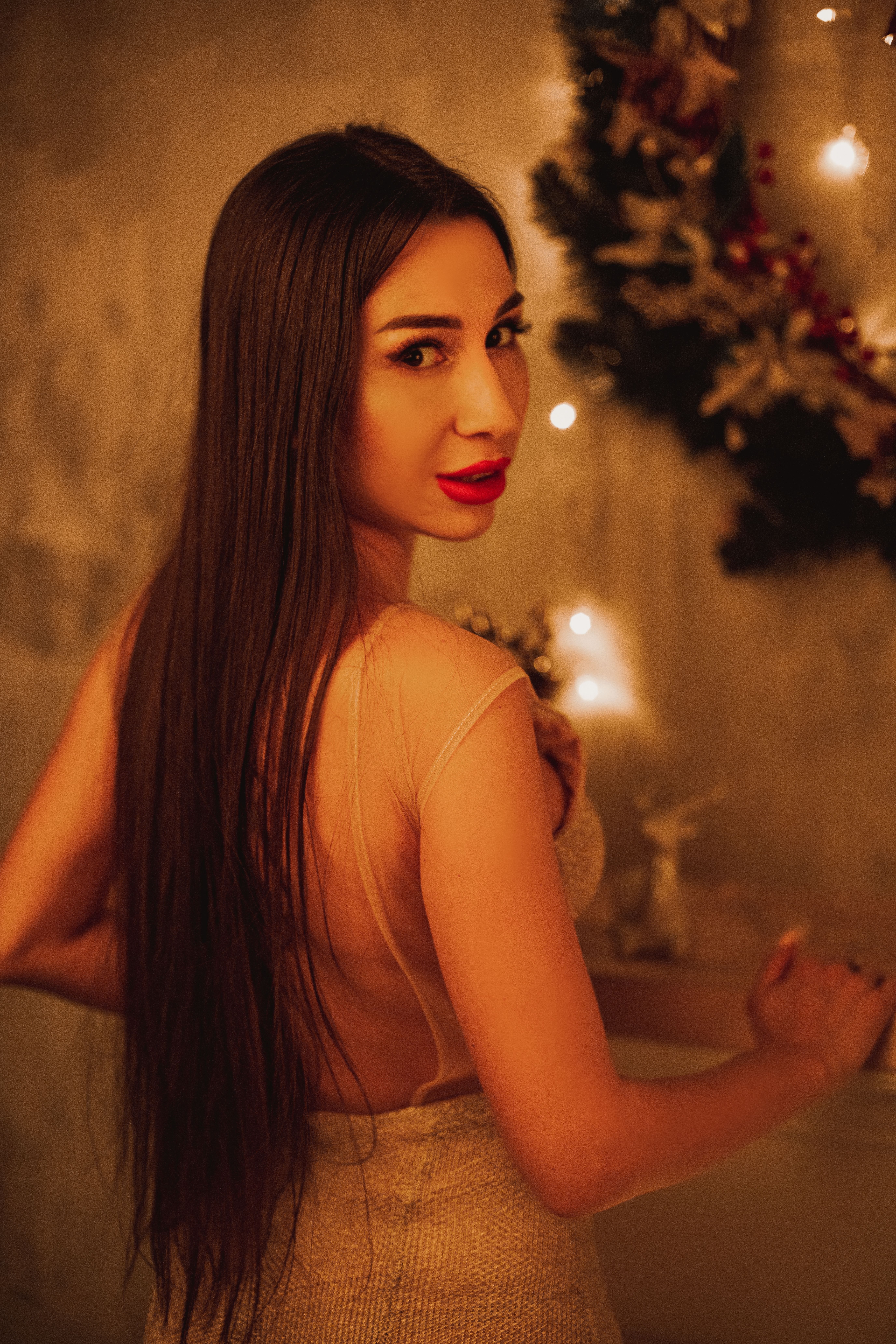 Stunning woman with long hair in a warm, festive indoor setting, looking over her shoulder.