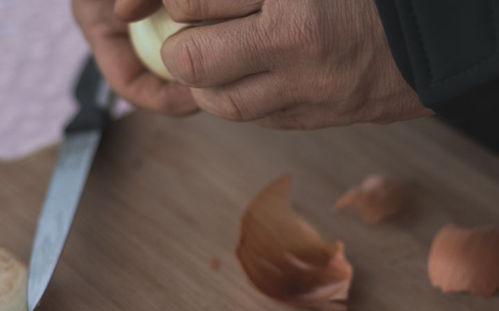 Detailed view of hands peeling an onion on a wooden cutting board, showcasing food preparation.