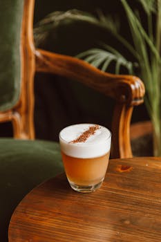 A beautifully crafted coffee with foam in a glass, placed on a wooden table near an antique chair indoors.
