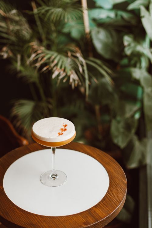 Free A sophisticated cocktail served in a glass with a foamy top, set on a wooden table against lush greenery. Stock Photo
