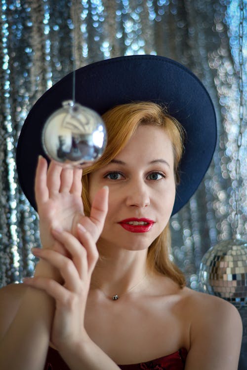 Gorgeous female in stylish hat looking at camera while touching hanging silver Christmas bauble in decorated room during festive event