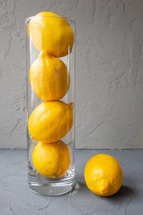 Glass vase with fresh whole lemons stacked on each other placed on table against gray wall