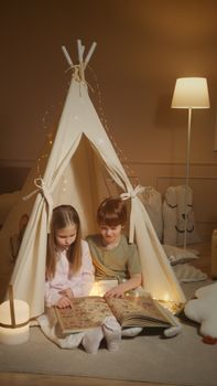 Two children enjoying a bedtime story inside a cozy indoor tent adorned with string lights.