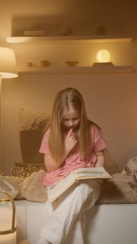A young girl in a pink dress reads a storybook in a cozy bedroom setting with warm lighting.