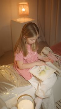 A girl sits reading a book in a softly lit, cozy bedroom environment.