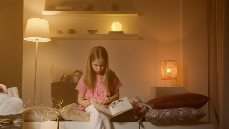 Girl In Pink Pajamas Reading A Book With Warm Lights Inside The Bedroom