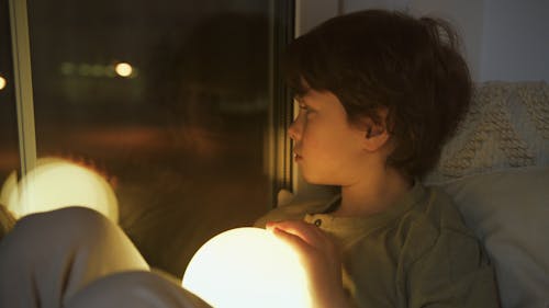 Boy Holding A Glowing Lamp Looking Through A Window 
at Night