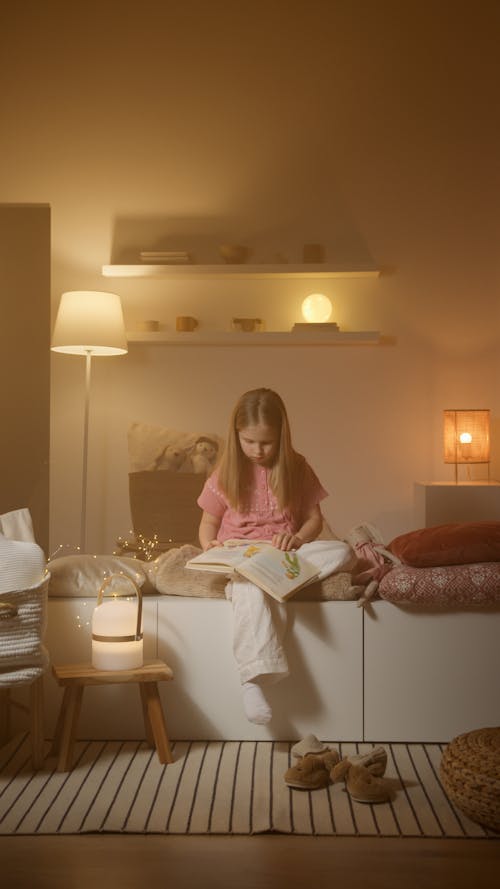 Free Girl In Pink Pajamas Reading A Book With Warm Lights Inside The Bedroom Stock Photo