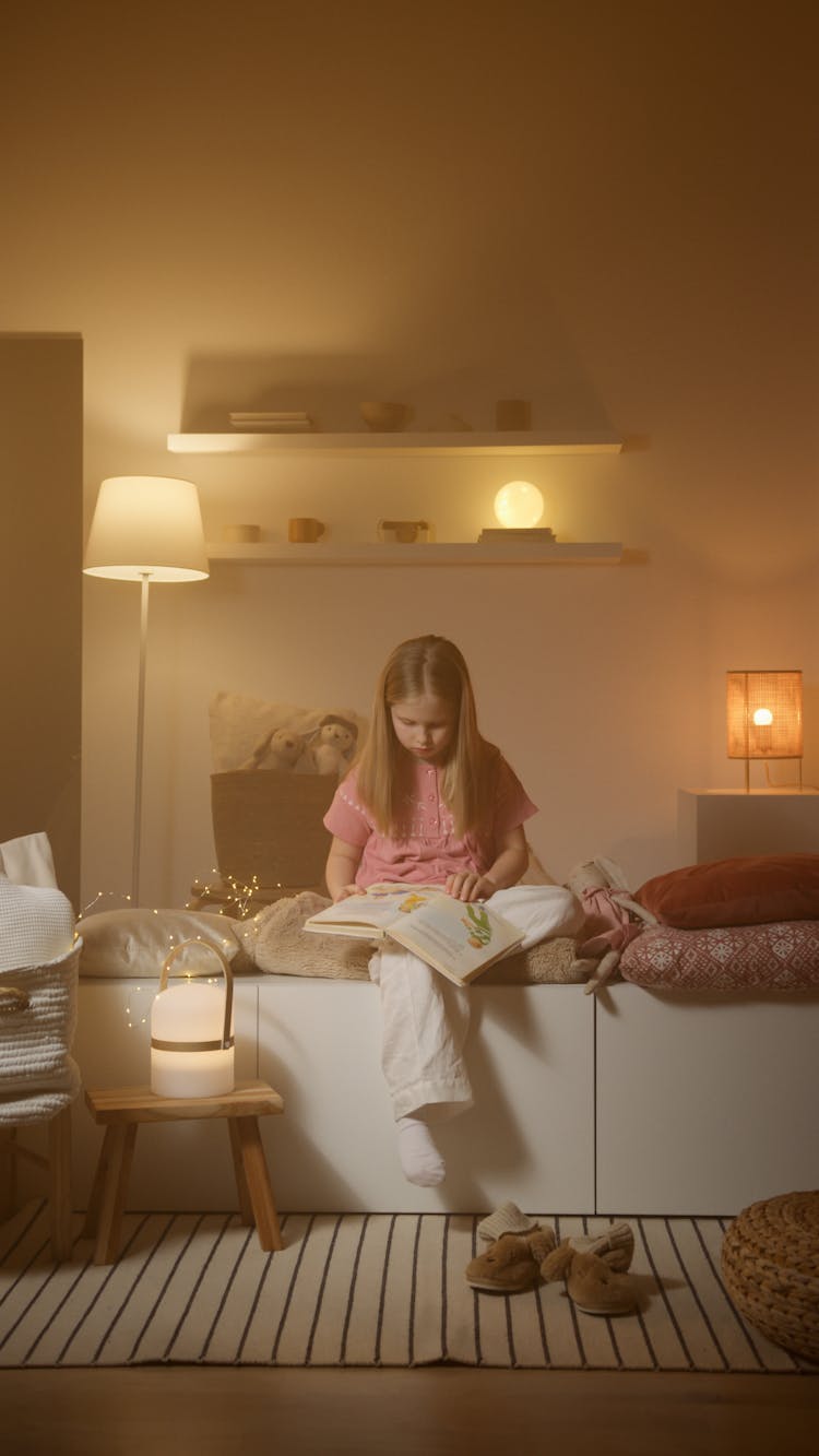 Girl In Pink Pajamas Reading A Book With Warm Lights Inside The Bedroom