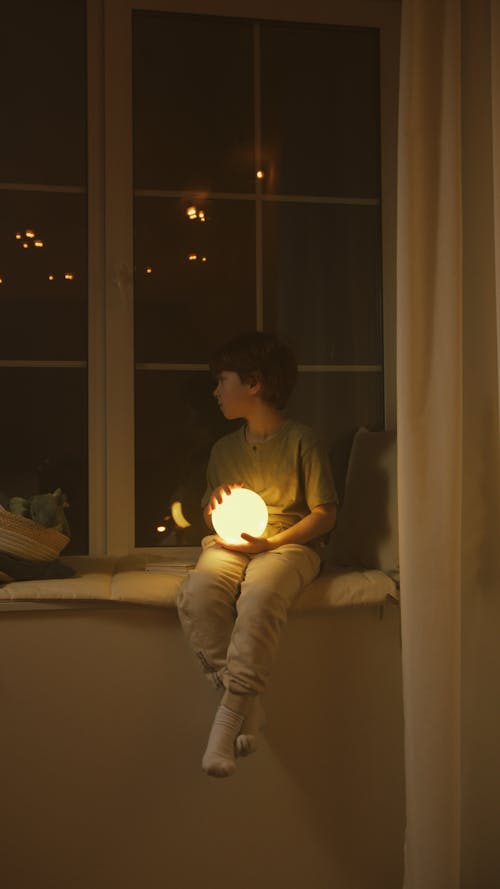 Free A Boy Holding a Luminous Ball while Sitting on the Windowsill Stock Photo