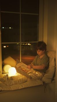 Child in pajamas reading on a window seat by warm glowing light. Cozy, serene night atmosphere.