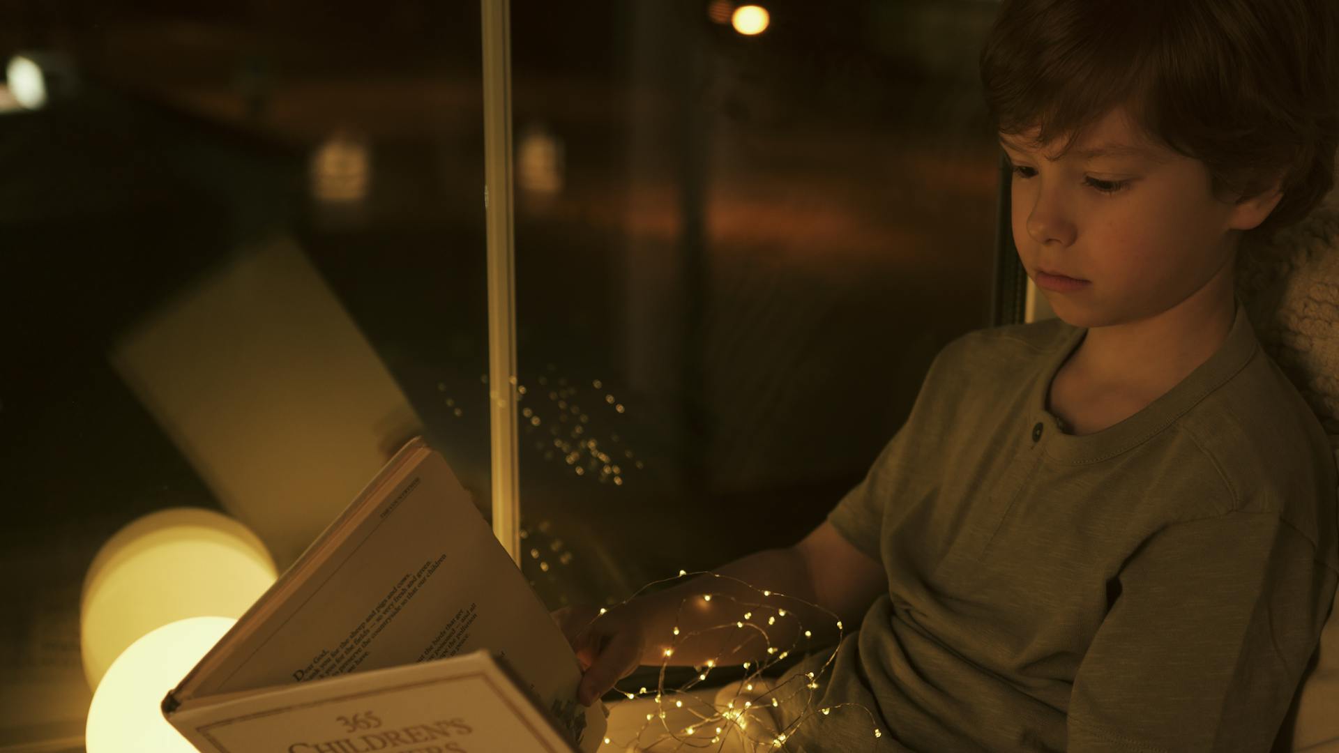 A young boy reads a children's storybook next to a window, illuminated by warm lights.