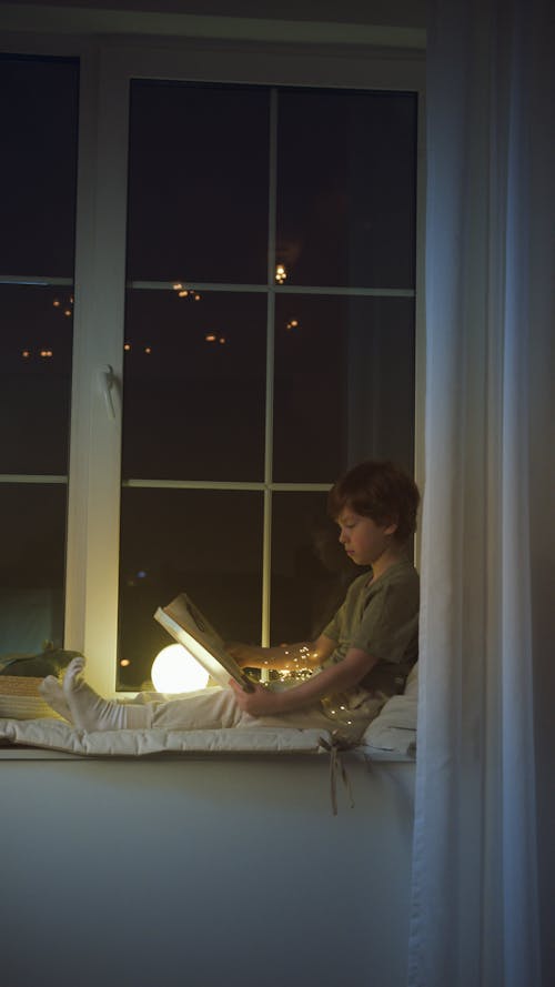 Free A Boy Reading a Book while Sitting on the Windowsill Stock Photo