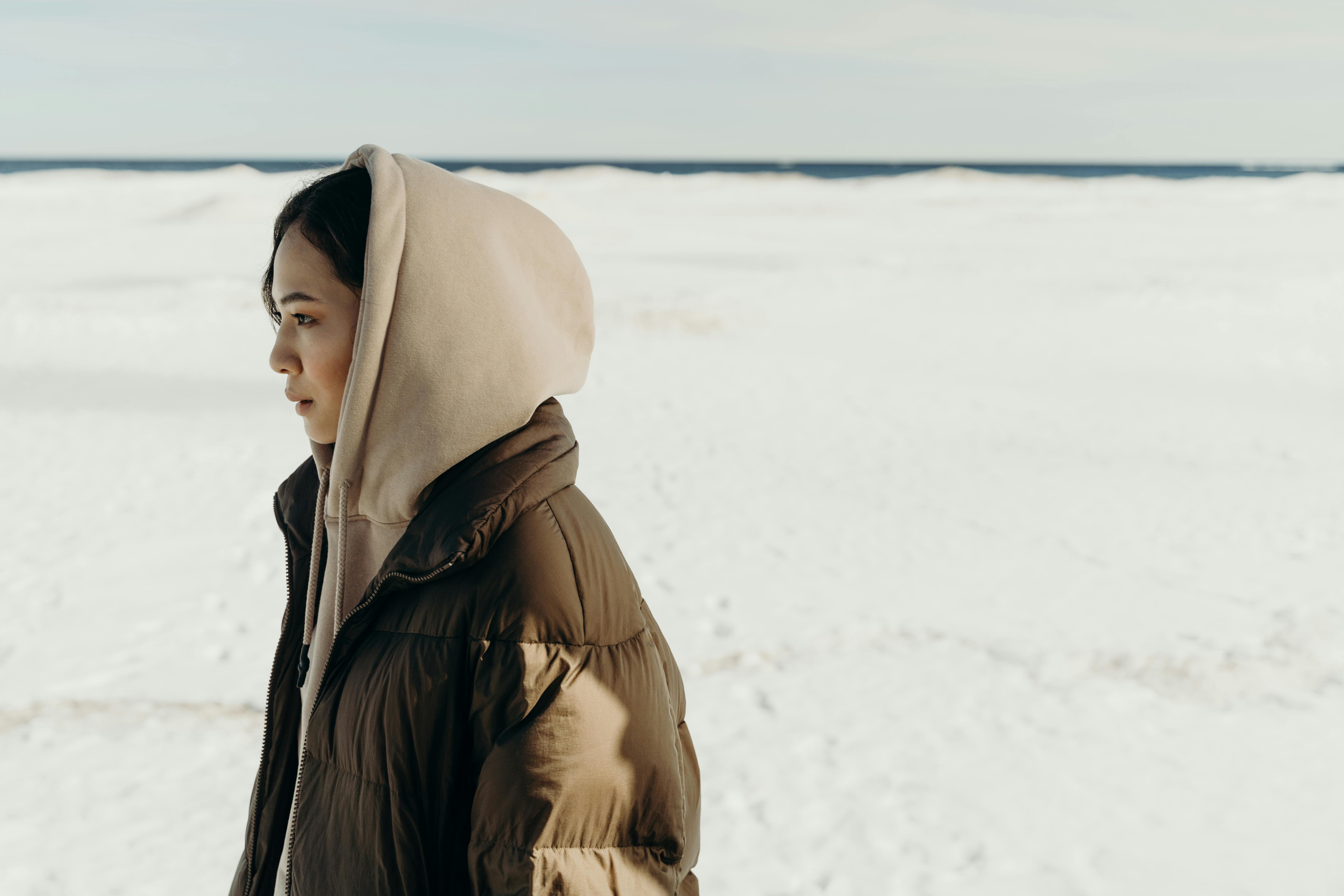 Side view of an Asian woman in a hooded puffer jacket walking through a snowy landscape.