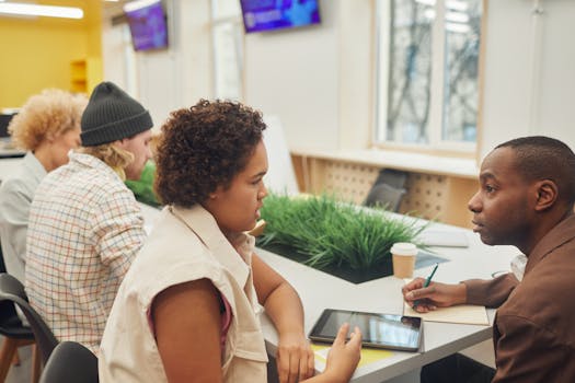 A diverse group of adults discussing work in a modern office setting with digital devices and green decor.