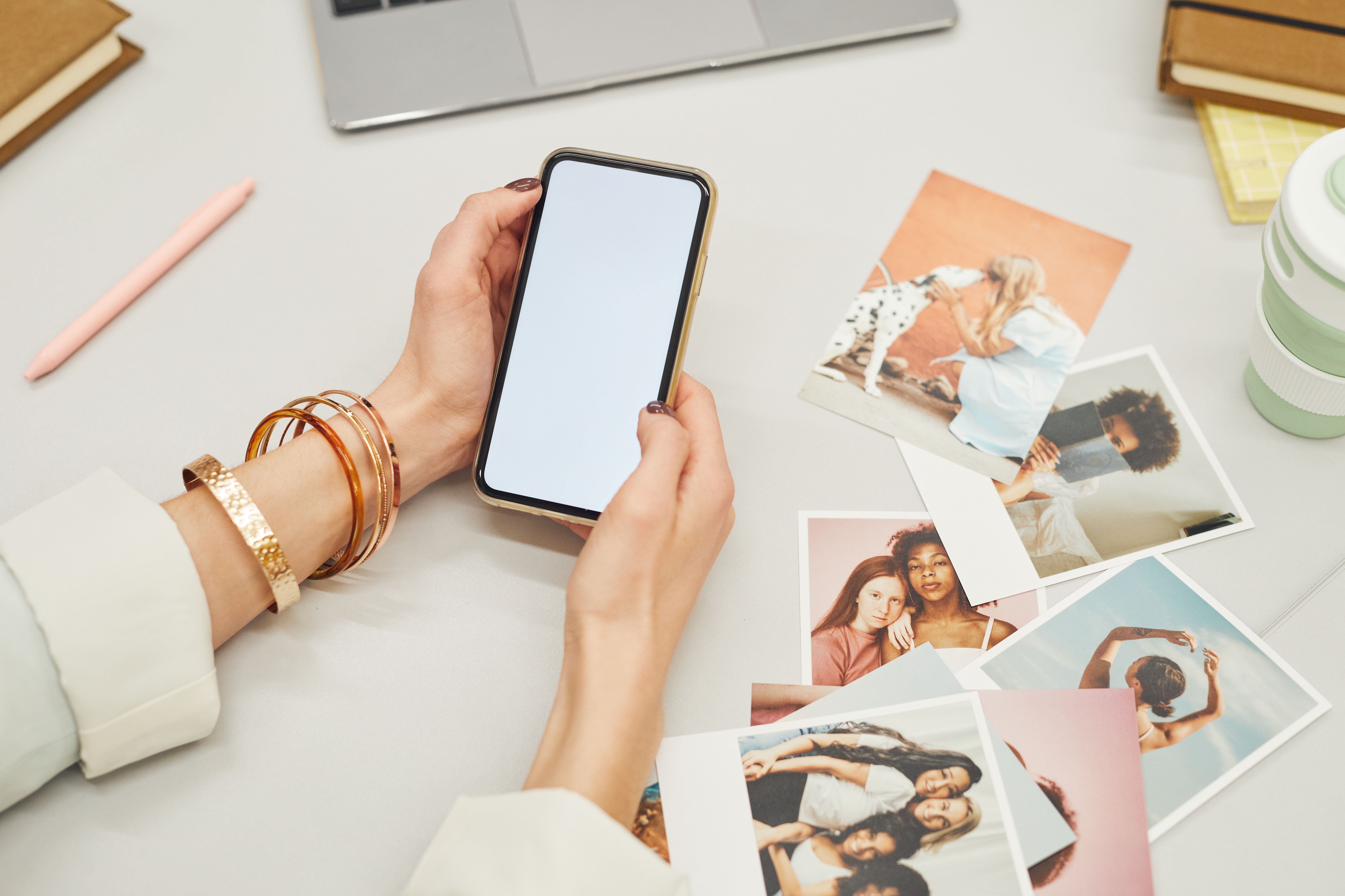 A person holding a smartphone surrounded by printed photographs and a laptop on a desk.