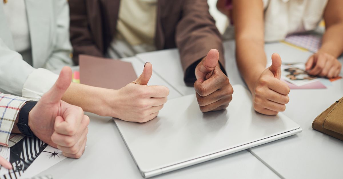 Four diverse hands showing thumbs up over a laptop on a desk, symbolizing teamwork and approval.