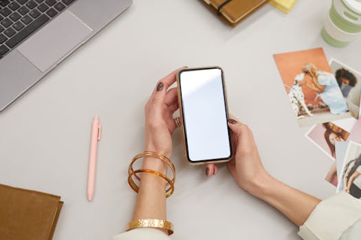 Close-up of a woman holding a smartphone on a desk with a laptop and photos, capturing modern workspace vibes.