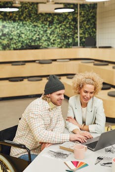 Two colleagues collaborating in a modern office space with a laptop and creative materials.