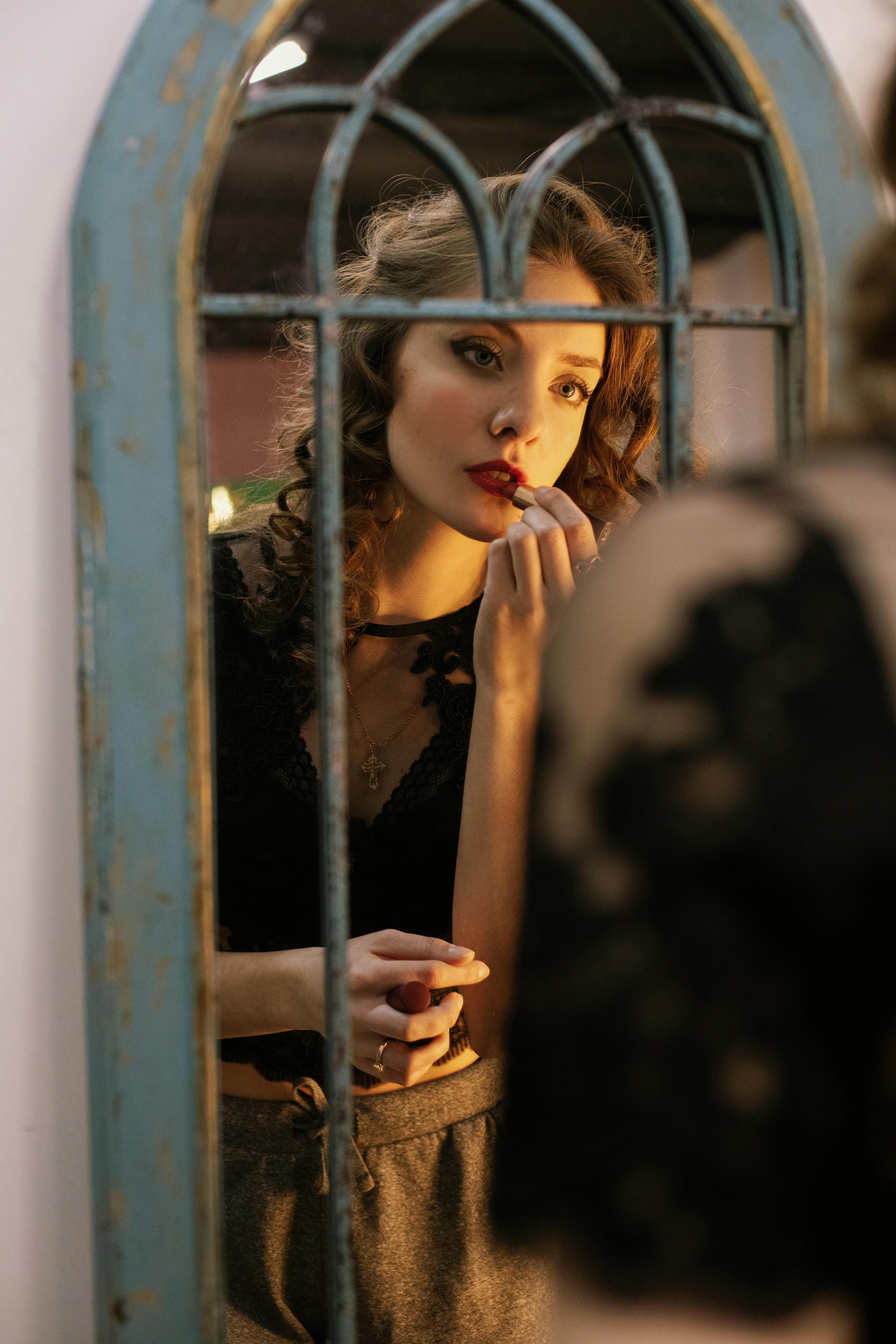 A young woman applies lipstick while looking into a vintage mirror, showcasing a classic beauty routine.
