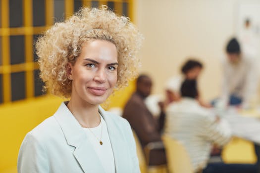 Portrait of a smiling businesswoman in blazer at a team meeting, with colleagues in the background.