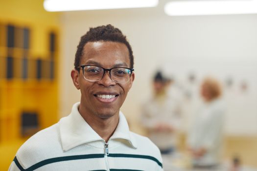 A smiling man wearing glasses in a cozy, well-lit indoor space with blurred figures in the background.