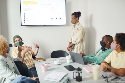 A diverse team discusses a project in an office setting with face masks, maintaining pandemic protocols.