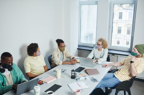 Free Office meeting with diverse colleagues wearing face masks, discussing projects in a modern workspace. Stock Photo