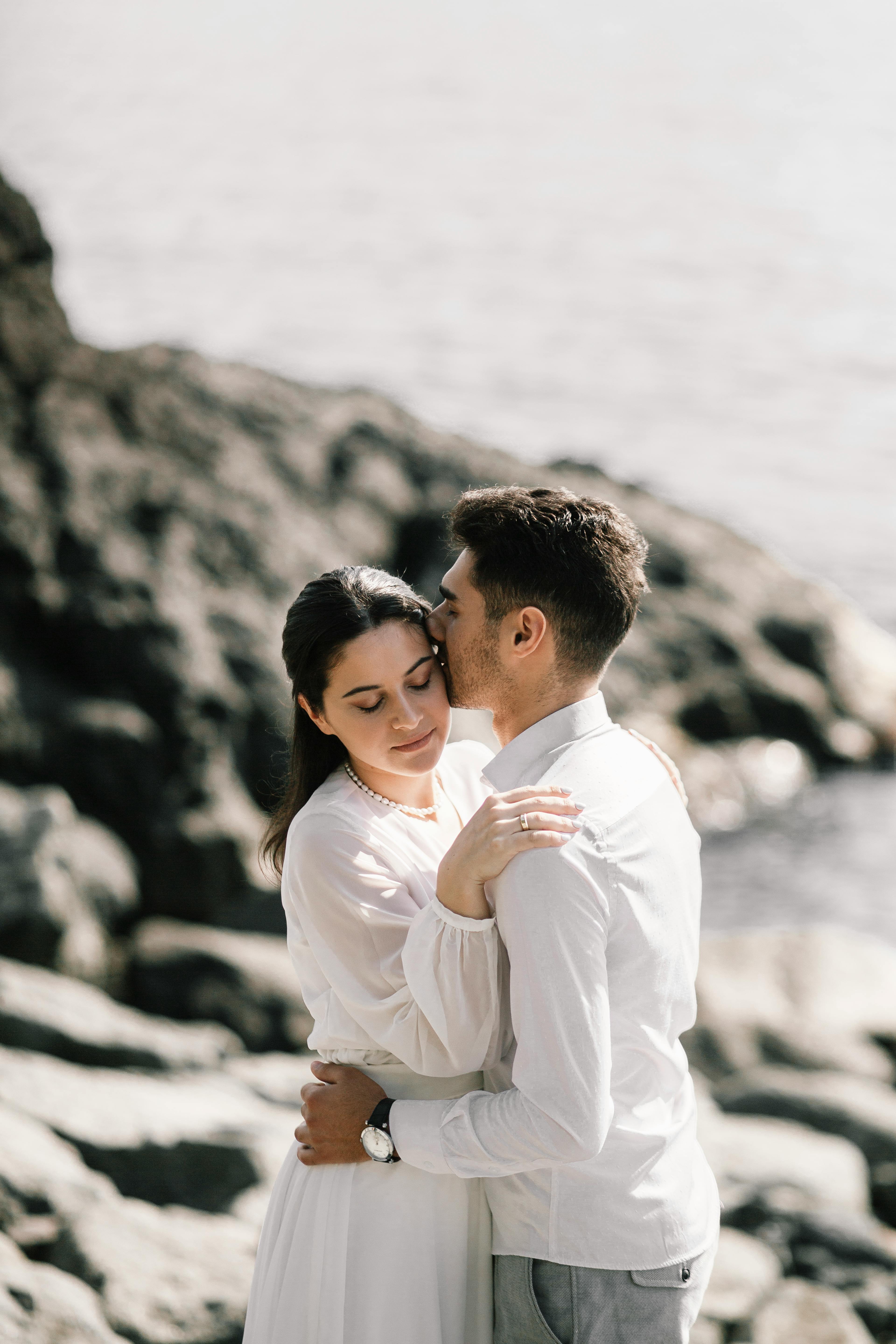 A romantic couple in white embraces on rocky seaside cliffs with a serene atmosphere.