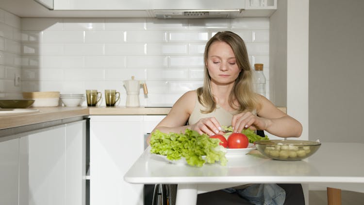 A Woman In Beige Tank Top Sitting On The Kitchen Near The Table With Vegetables