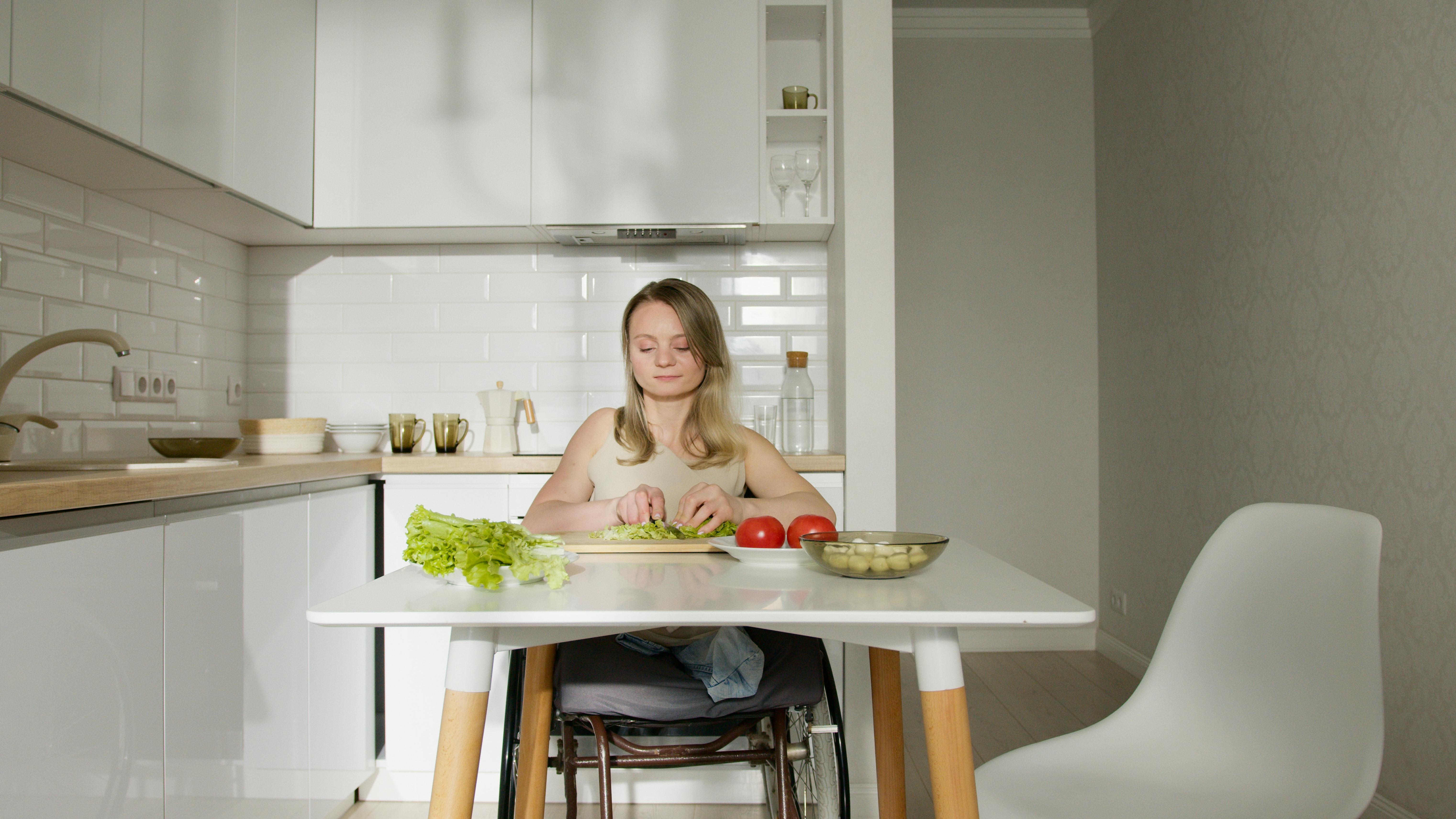 Free Young woman in a wheelchair slicing vegetables at a stylish kitchen table. Stock Photo