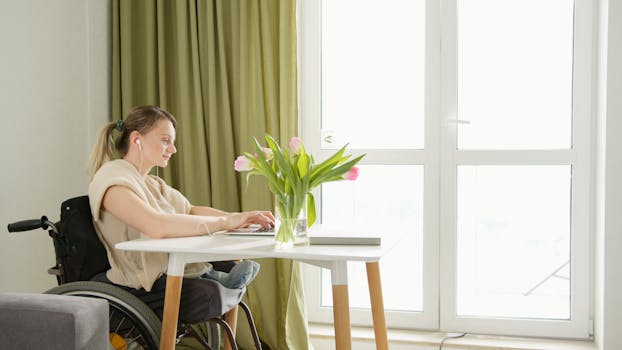 A woman in a wheelchair working at a home office desk with a laptop and flowers.