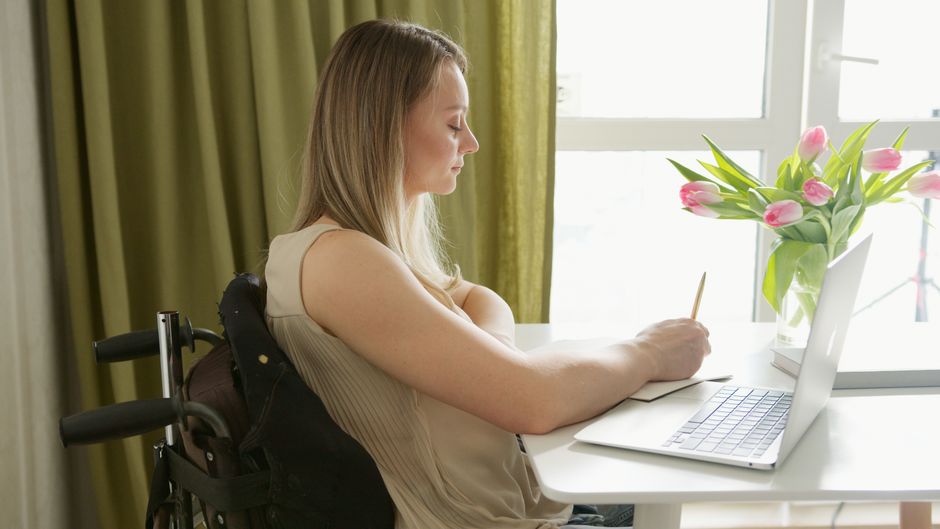 Mujer en silla de ruedas trabajando en un portátil en casa cerca de un ramo de flores.