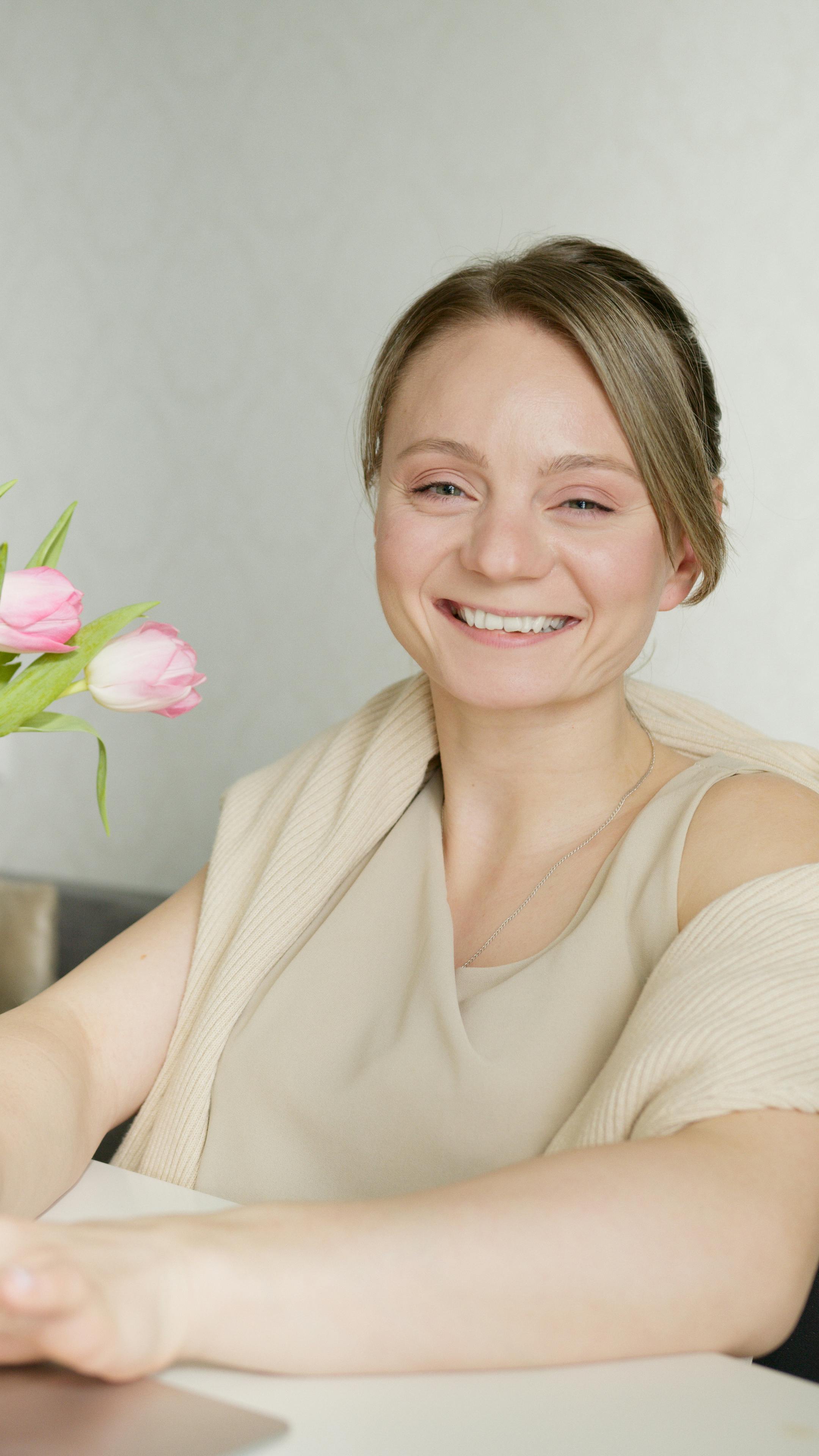 A joyful woman smiling indoors beside a vase of pink tulips, exuding a warm and serene atmosphere.