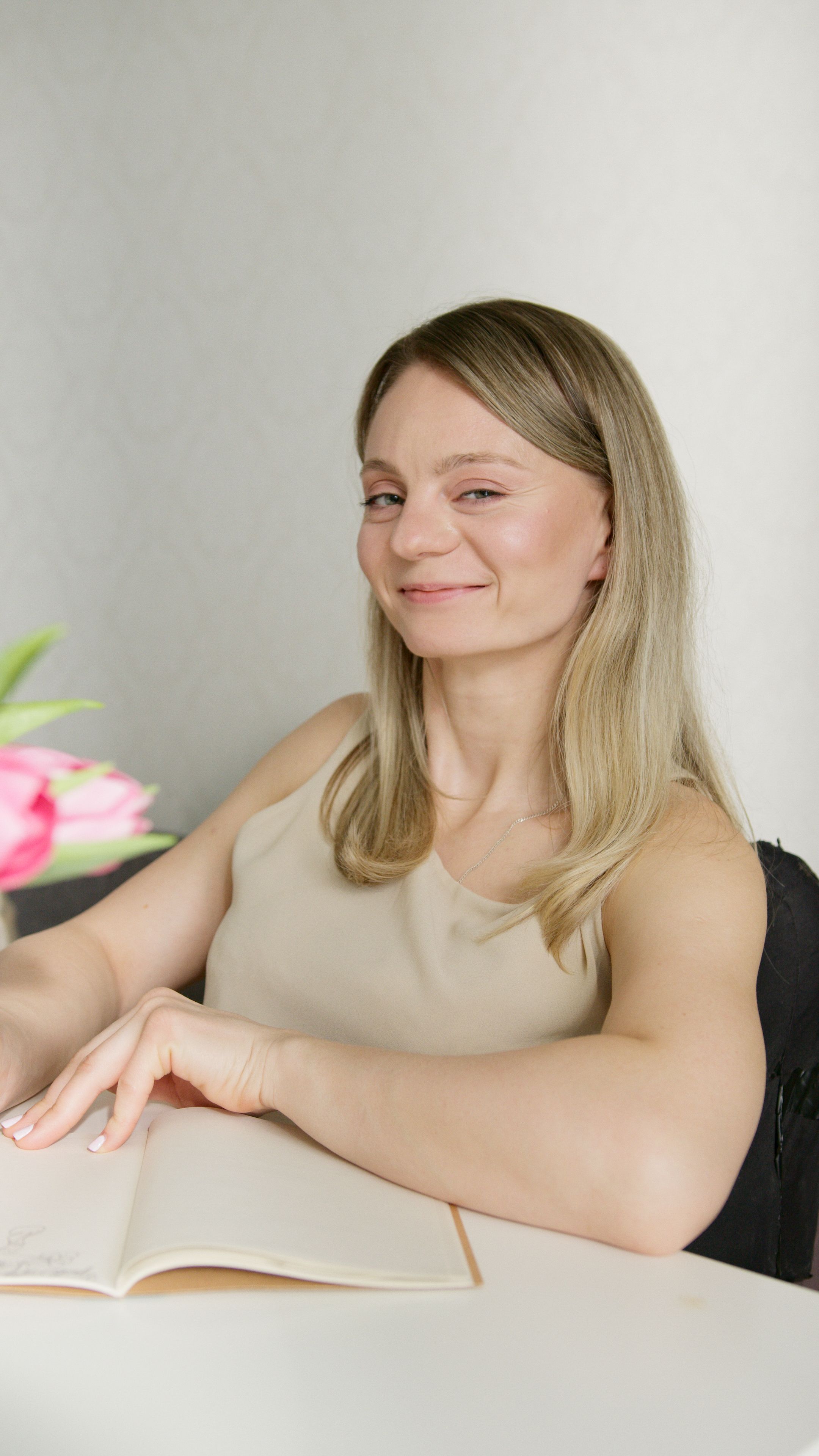 Young woman with blonde hair sitting indoors, smiling while holding a notebook on a table.
