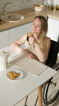 A woman in a wheelchair relaxes with coffee in a bright kitchen, embracing independence.