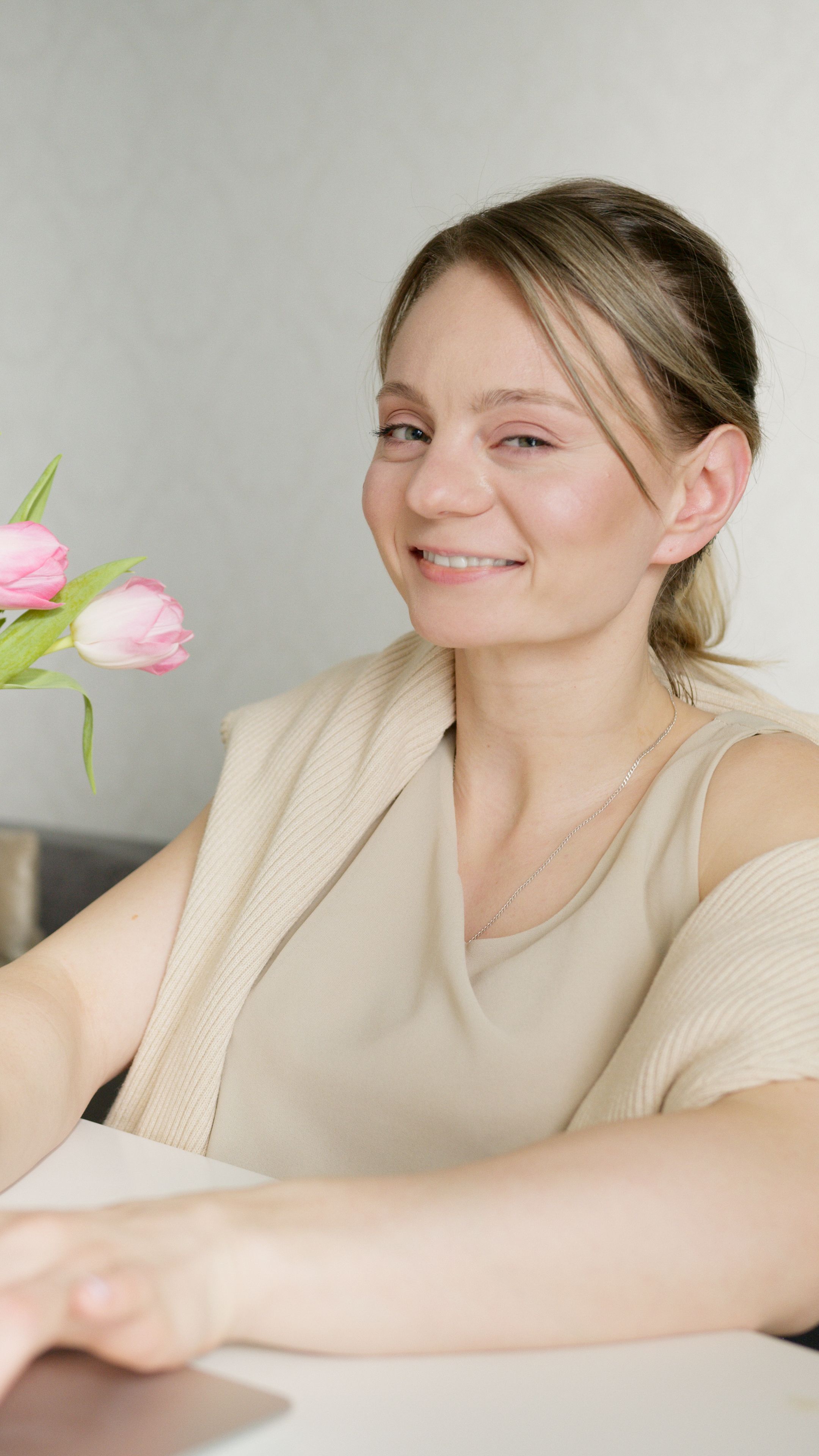 A cheerful woman smiles while seated at a table with pink tulips, in a bright and cozy indoor setting.