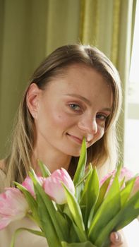 A close-up portrait of a smiling woman holding pink tulips indoors with natural light.