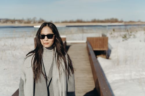 Free Asian woman in winter attire standing on a snowy boardwalk, enjoying the serene countryside view. Stock Photo