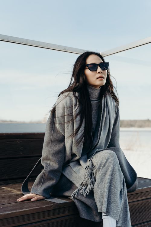 Free Elegant woman in gray coat and sunglasses sitting on a wooden bench, showcasing modern style. Stock Photo