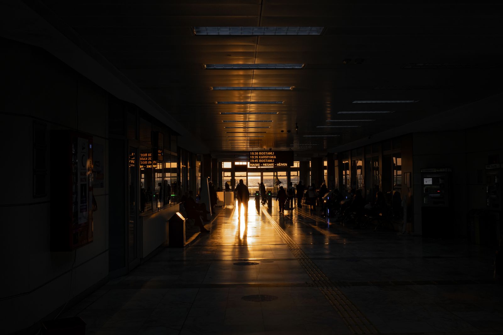 Travelers silhouetted walking through a dark airport terminal as warm sunset light streams across the floor