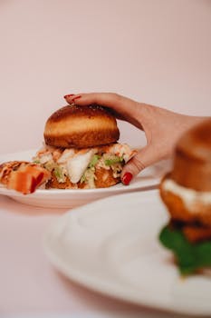 A gourmet lobster roll in a bun, elegantly plated, held by a hand with red nails.