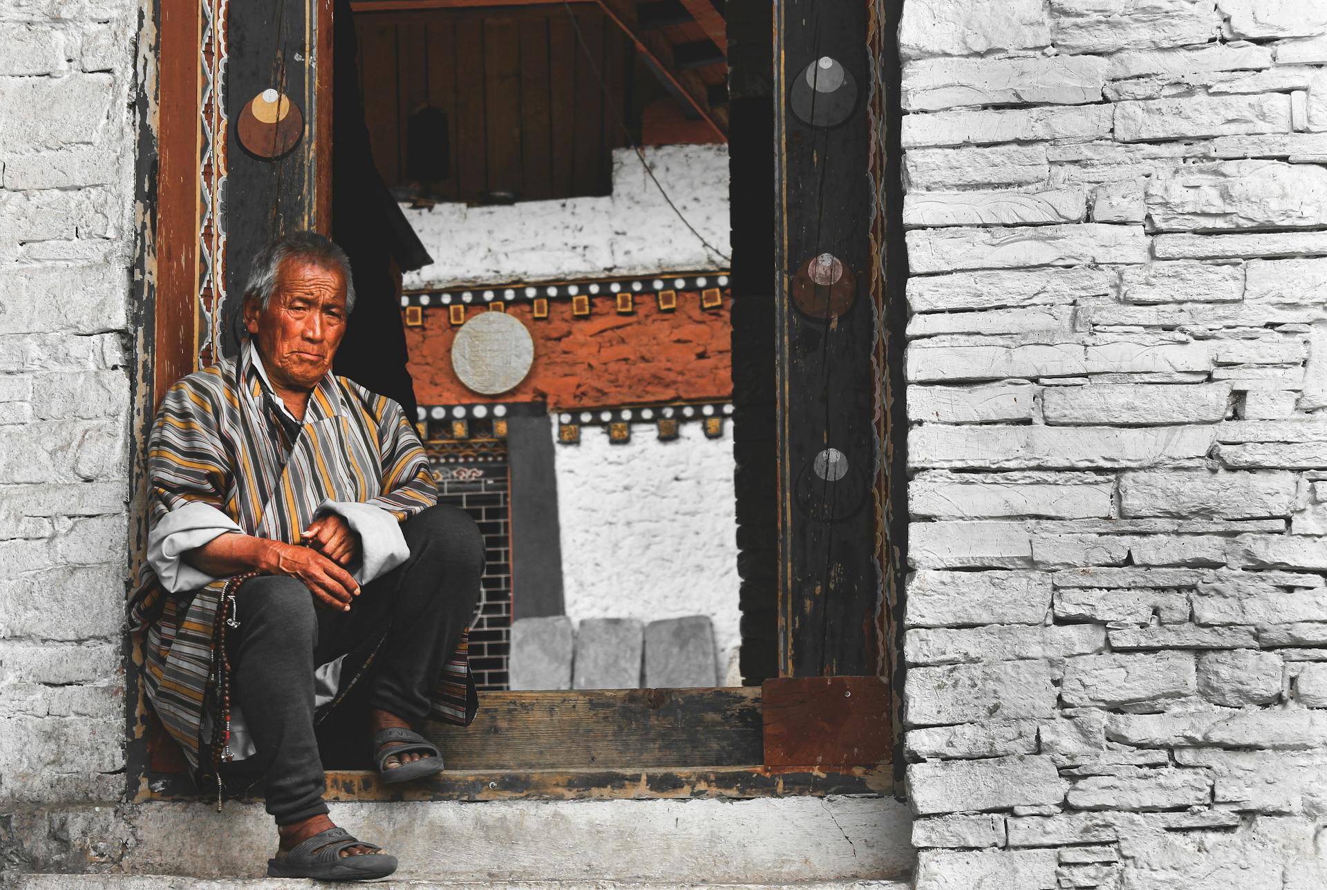 Elderly Bhutanese man in traditional attire sits in a monastery doorway, Bhutan.