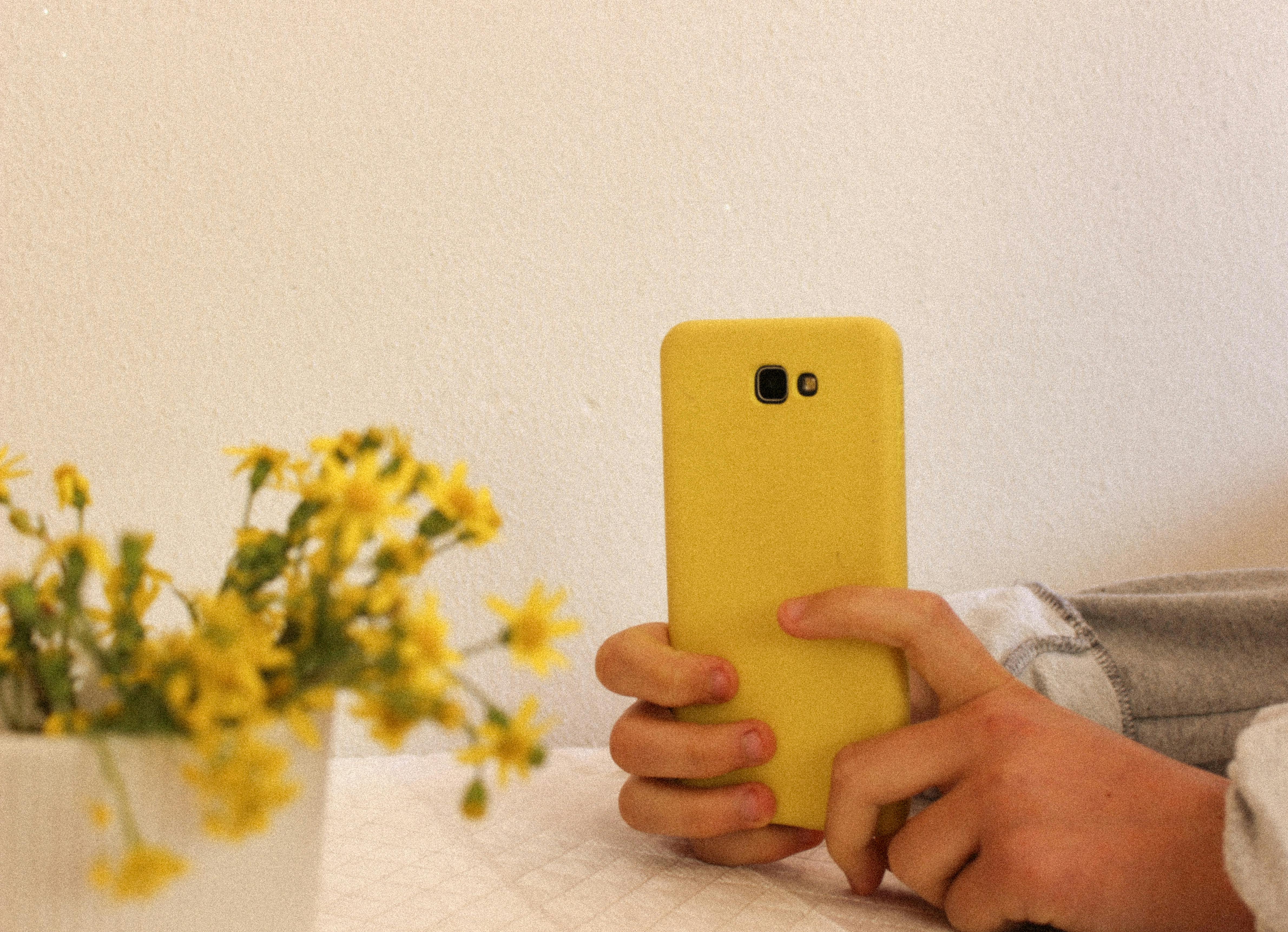 Hands holding a smartphone with a yellow case next to a vase of flowers indoors.